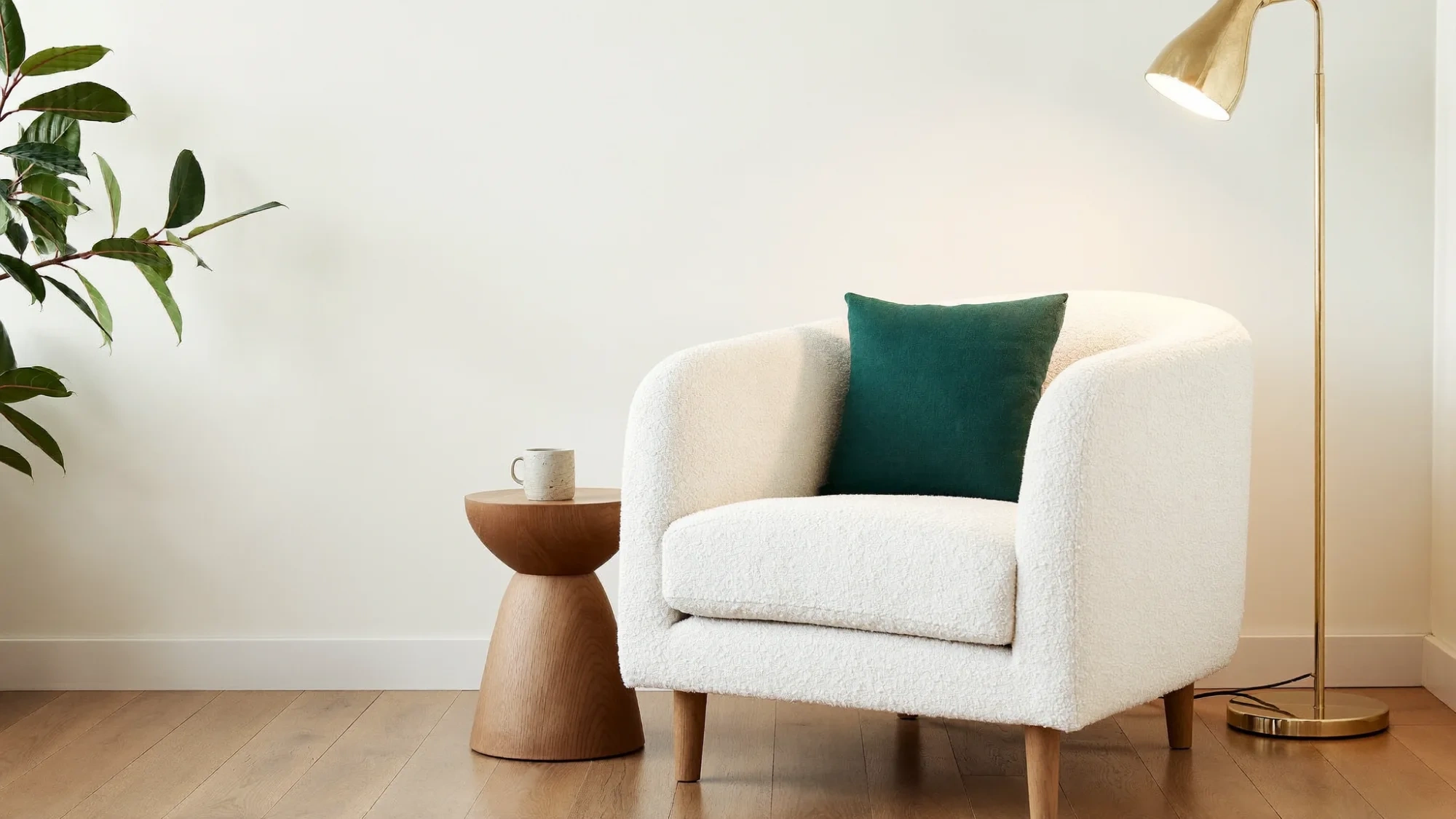 A cream bouclé armchair with a deep forest-green cushion beside a brass floor lamp and oak side table, in bright morning light against a clean cream wall