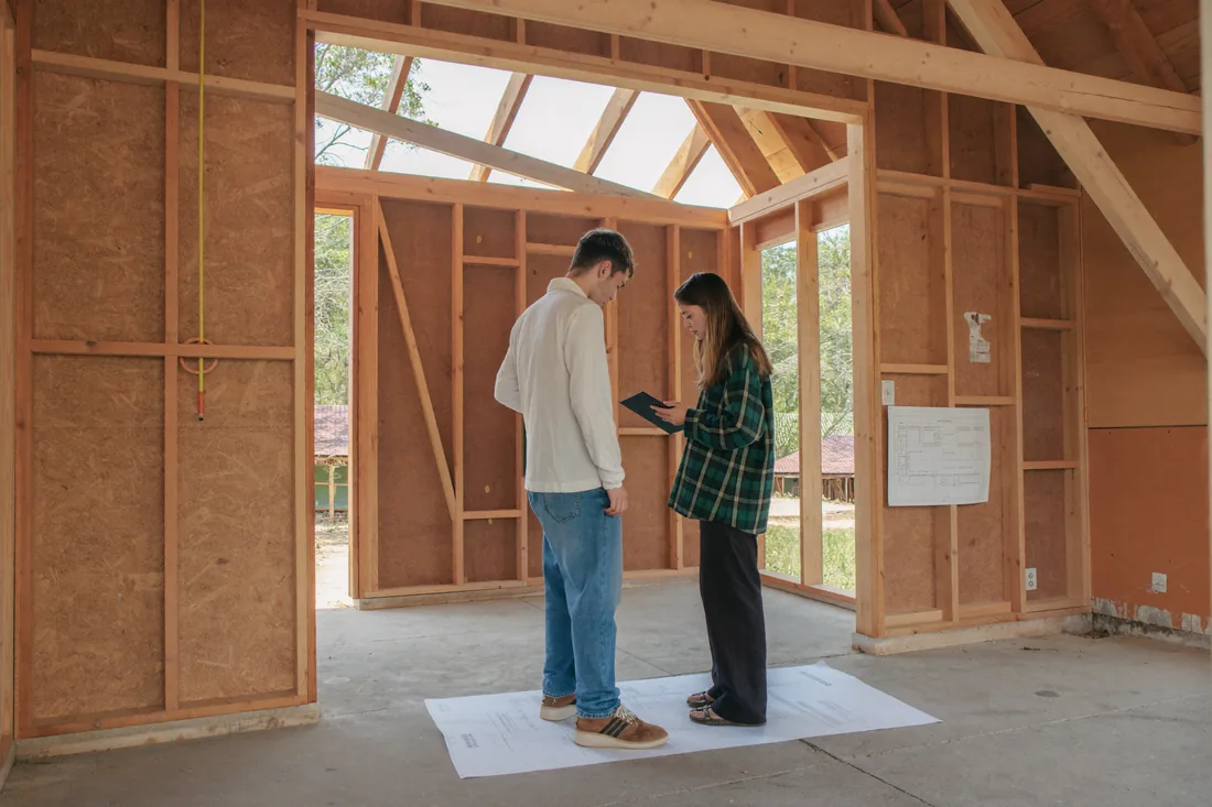 A young Australian couple standing inside the timber frame of their new home during construction, reviewing building plans on the site.
