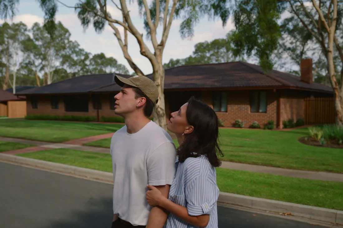 A young couple standing outside a brick three-bedroom house in a Canberra suburb, the kind of first home bought on a 99-year ACT Crown lease.