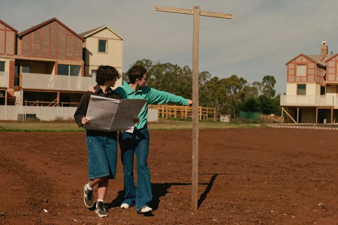 A young couple inspecting a vacant suburban building block in a new Australian estate, checking the boundary against a site plan with half-built homes nearby.