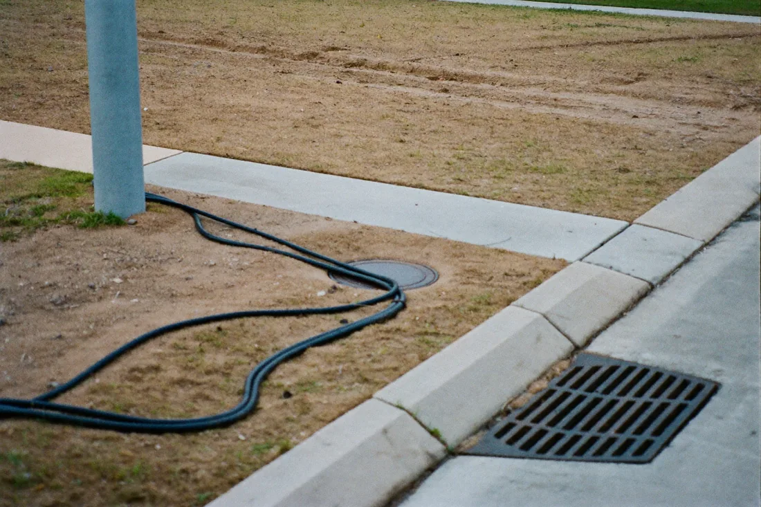 Utility service connections at the boundary of an Australian vacant residential lot, a kerbside power pole, a stormwater drainage access lid, and an NBN utility pit.