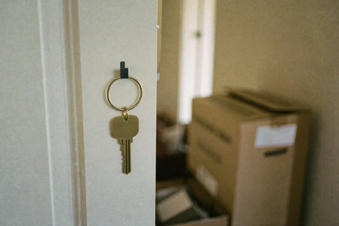 A single brass key hanging from a small hook on a door frame, with stacked cardboard moving boxes out of focus in the sunlit room behind.