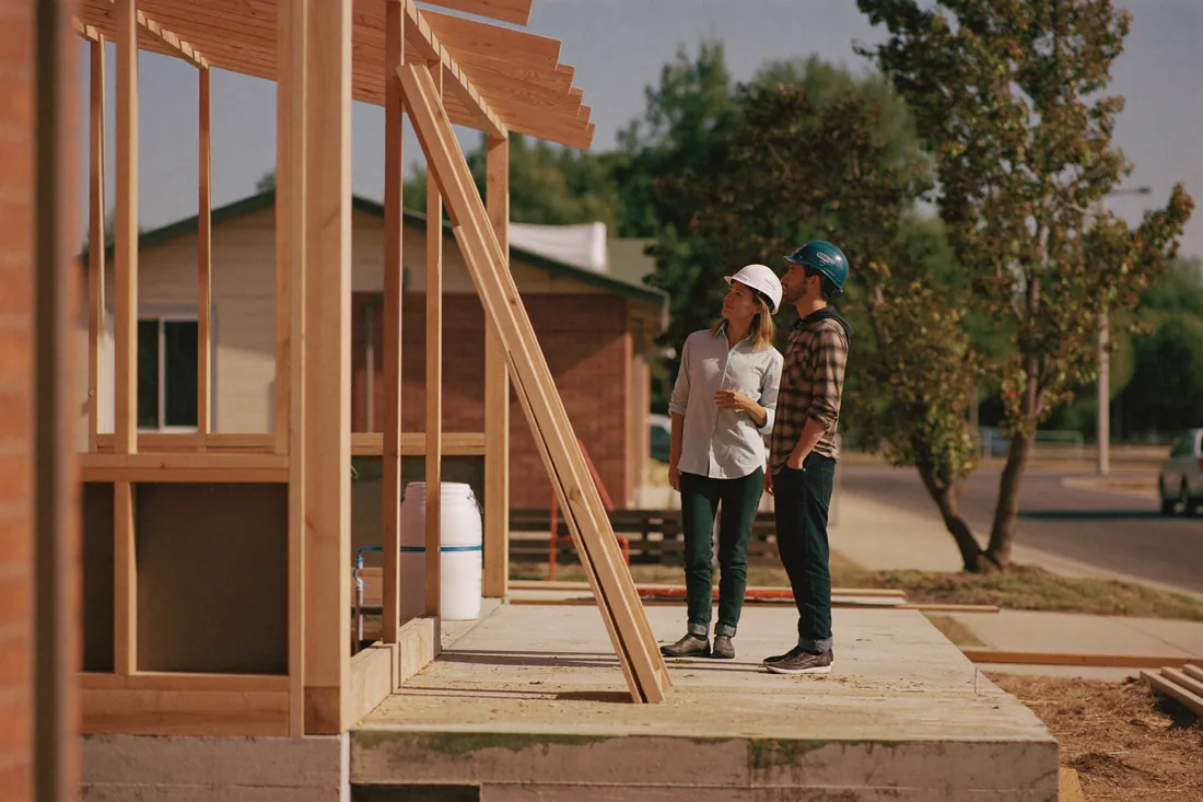 A young Australian couple stand on the concrete slab of their new home as the timber wall frame and roof trusses go up during the lock-up stage of construction.