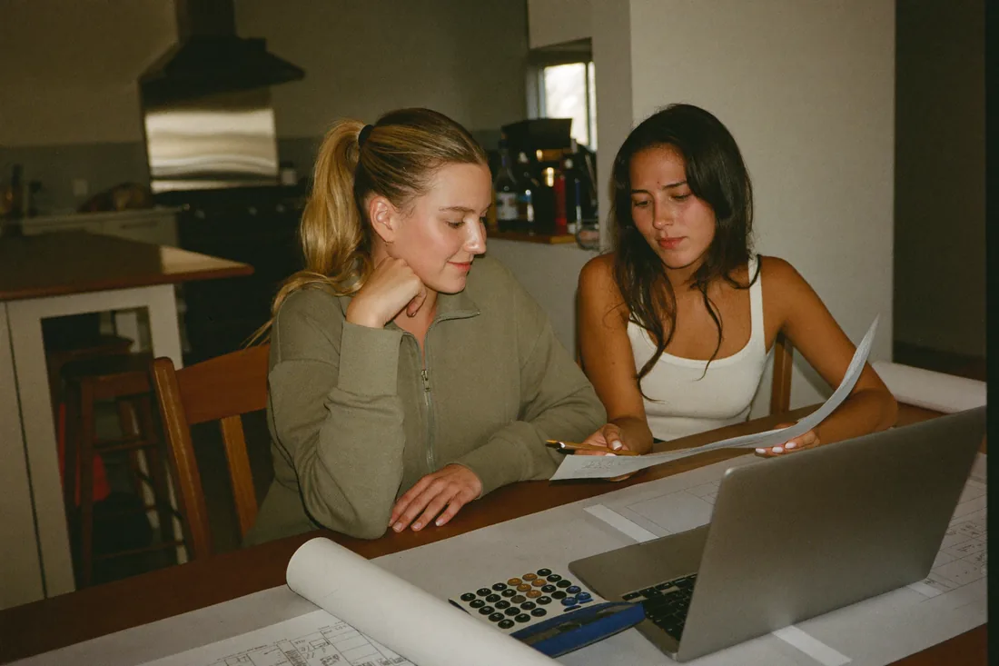 Two first home buyers sit at a kitchen table comparing their building contract, grant paperwork and deposit on a laptop while planning a new-build construction loan.