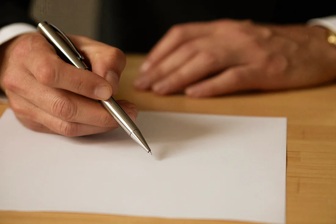 A close-up of a hand holding a silver pen over a blank page, with a second hand resting flat on the table