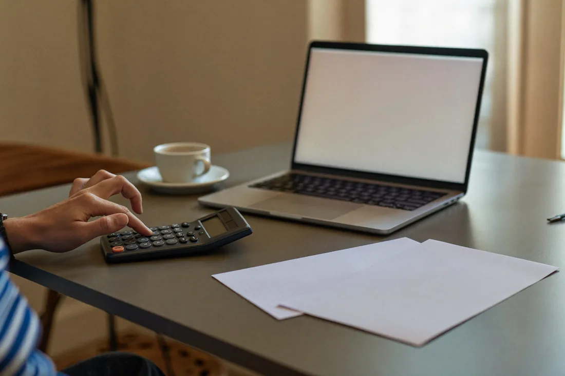 A person at a kitchen table using a calculator and laptop to work out the yearly cost of owning their home.