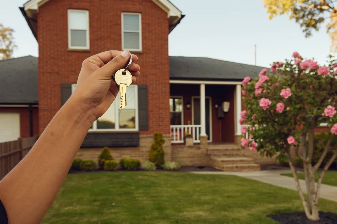 A hand holding the house keys in front of an established brick home, the point at which a buyer is finally safe from being gazumped.