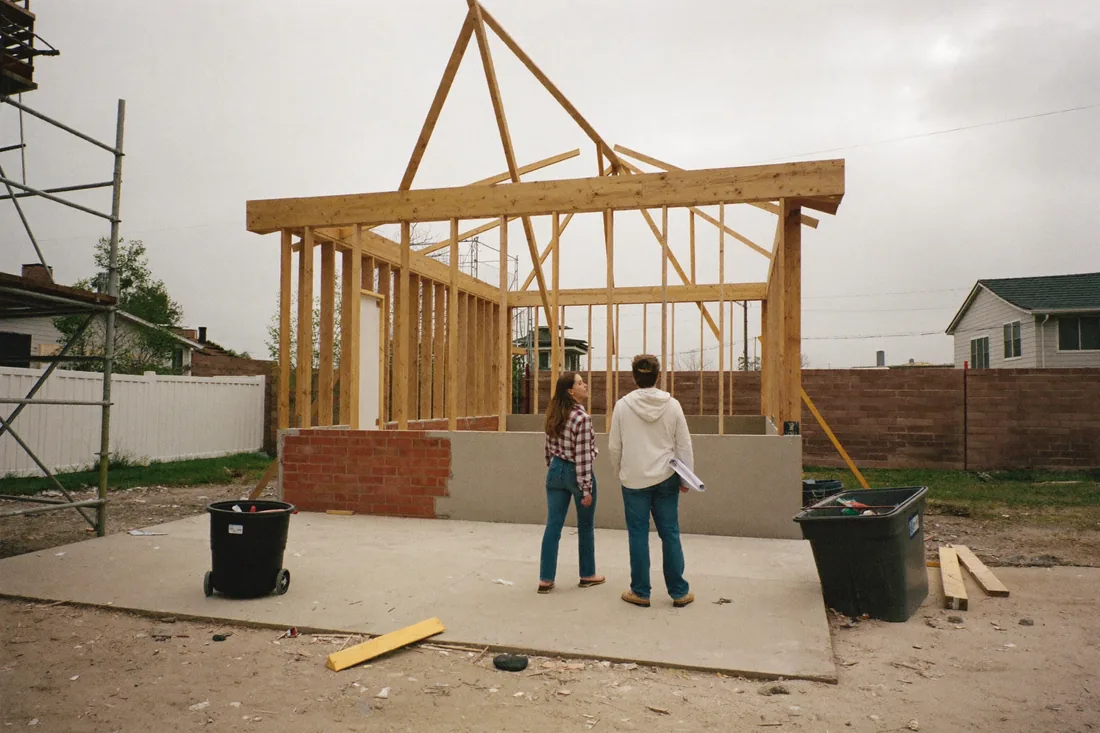 A first-home-buyer couple standing on the concrete slab of their half-built home, timber frame and roof trusses up, on a suburban Australian block.