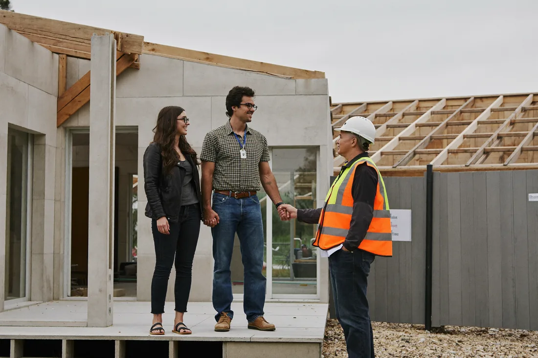 A young couple talking with a site supervisor in a hi-vis vest and hard hat on the slab of their partly-built new home on a suburban Australian block.