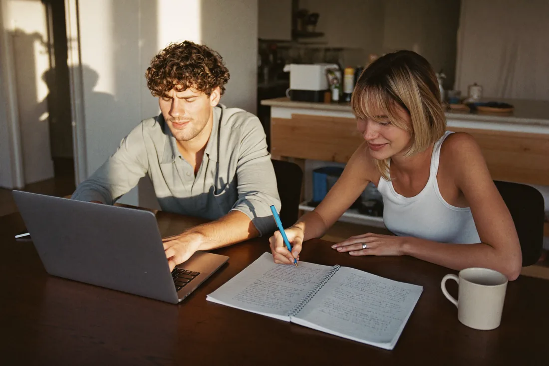 A young Australian couple working out their house deposit at the kitchen table with a laptop and notepad.