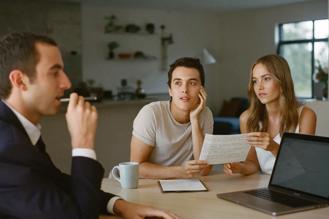 A first-home-buyer couple interviewing a mortgage broker at their kitchen table, reading from a printed list of questions.