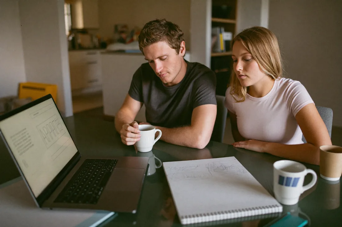 A young couple at their kitchen table working out their maximum auction bid on a laptop and notepad before auction day.