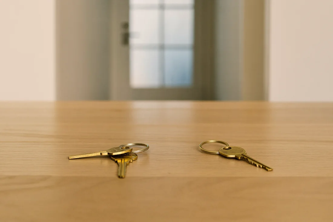 Two separate sets of brass house keys side by side on a timber table, with a blurred doorway behind