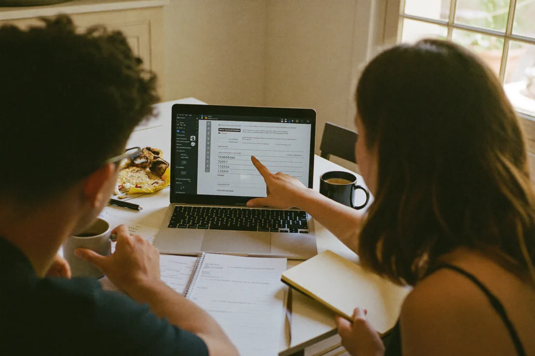 A young Australian couple at their kitchen table working through property numbers on a laptop while weighing renting versus buying.