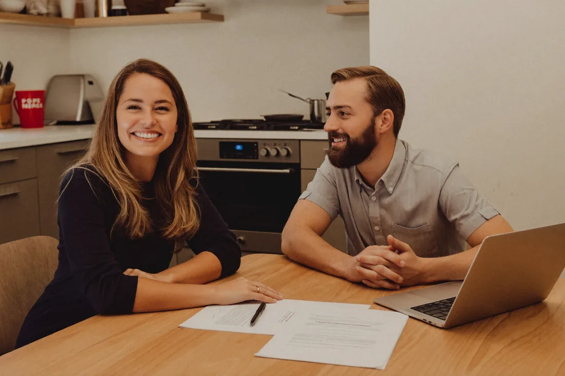 An Australian home seller and a real estate agent discussing the commission rate and agency agreement at a kitchen table, with a 'for sale' campaign brochure between them.